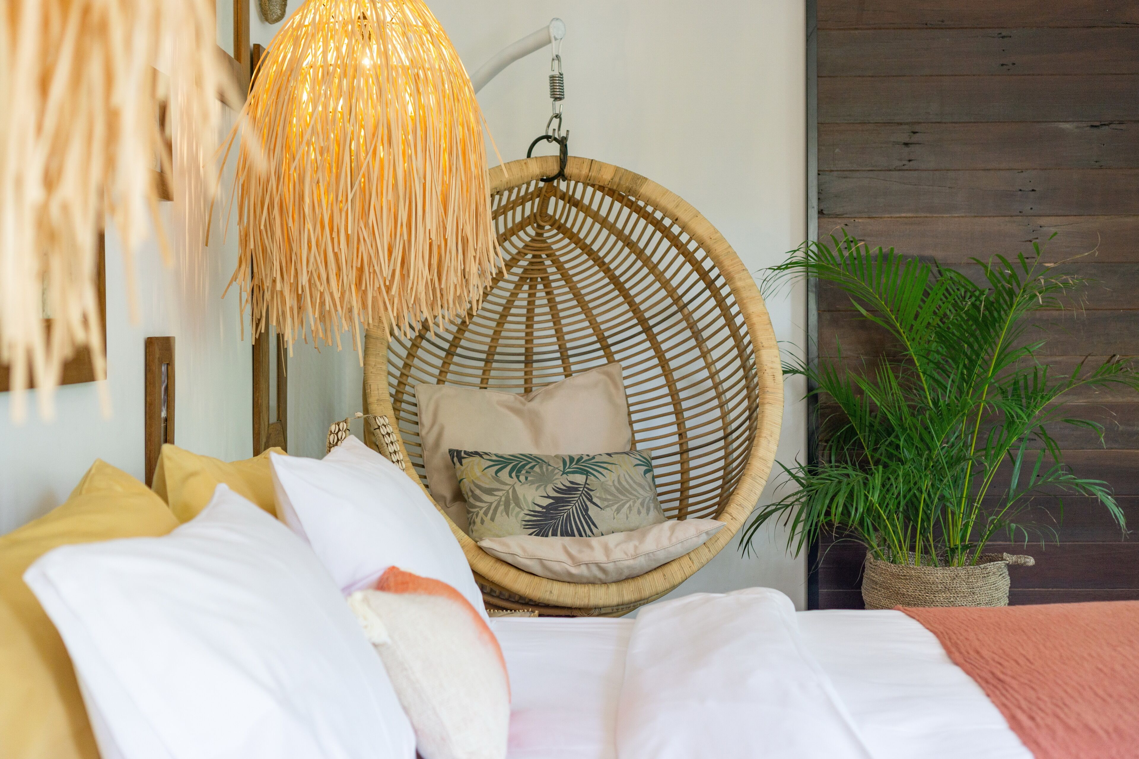 A bed with white and yellow bedding, a wicker chair with cushions, a hanging light fixture, and a potted plant.