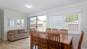 A room with a wooden dining table, four chairs, a beige sofa, and a large window with blinds. - Sandy Shoal, 46 Rigney Street - Shoal Bay Beach Cottage with aircon (Shoal Bay)