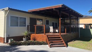 A single-story mobile home with a covered porch, a small patio area, and a front entrance. - Sandy Shoal, 46 Rigney Street - Shoal Bay Beach Cottage with aircon (Shoal Bay)