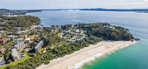 An aerial view of a coastal residential area with beachfront properties, dense vegetation, and a clear view of the ocean. - The Helm, 8/22 Voyager Close - hear the waves from Shoal Bay Beach (Nelson Bay)