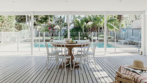 A covered outdoor area with a pool, white chairs, and a dining table.