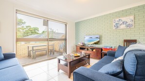 A modern living room with a large sliding glass door leading to an outdoor area, a flat-screen TV mounted on a brick wall, and a dining table with a plate of fruit.