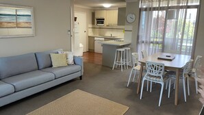 A compact living space with a sofa, dining table, chairs, a kitchen area, and a large window with curtains. - The Commodore, 14, 9-11 Donald Street- Unit in the heart of town with views & free wi-fi (Nelson Bay)