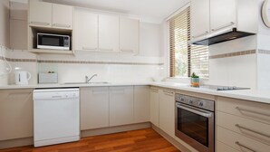 A modern kitchen with white cabinetry, a built-in microwave, a dishwasher, an oven, and a window with blinds. - The Commodore, 14, 9-11 Donald Street- Unit in the heart of town with views & free wi-fi (Nelson Bay)