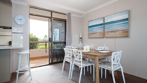 A dining area with a wooden table, white chairs, and a view through a glass door to the outdoors. - The Commodore, 14, 9-11 Donald Street- Unit in the heart of town with views & free wi-fi (Nelson Bay)