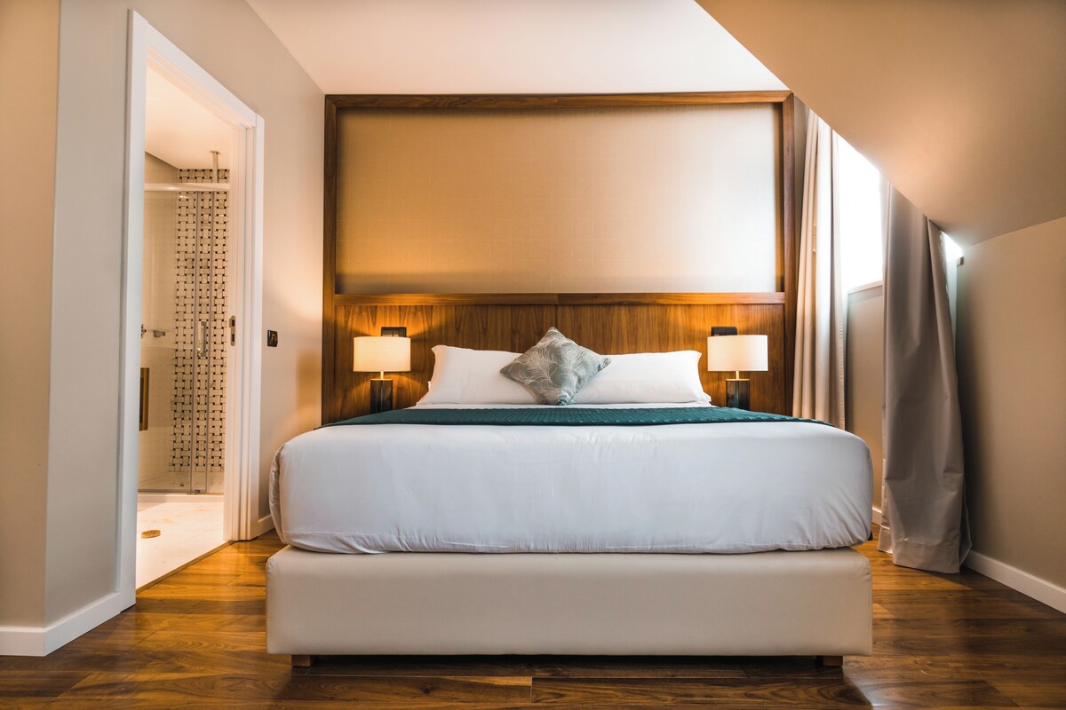 A hotel room with a large bed, two bedside lamps, a wooden headboard, and a view of the outdoors.