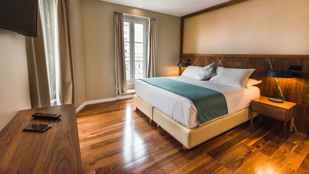 Ein Hotelzimmer mit einem großen Bett, einem an der Wand montierten Fernseher, einem Nachttisch mit Lampe und einem Fenster mit Vorhängen.
