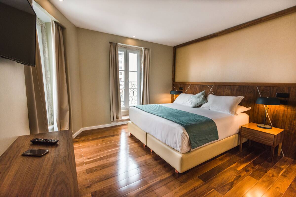Une chambre d’hôtel avec un grand lit, une table de chevet avec une lampe, un téléviseur fixé au mur et une fenêtre avec des rideaux.