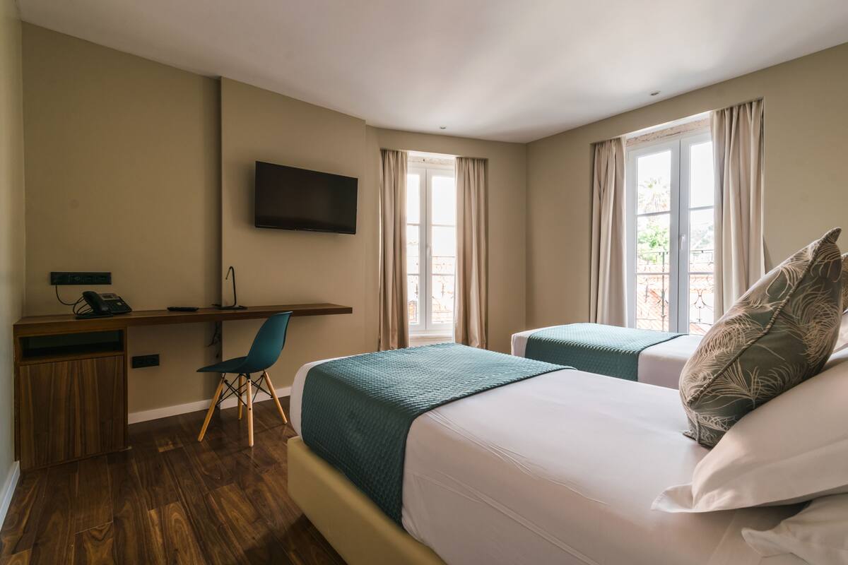 Une chambre d’hôtel avec deux lits, un bureau avec une chaise, un téléviseur fixé au mur et de grandes fenêtres avec des rideaux.