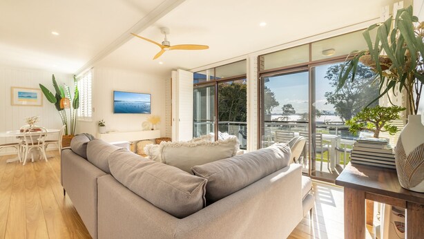 En moderne stue med en stor grå sofa, et spiseområde med et hvidt bord og stole, og en balkon med udsigt over havet.
