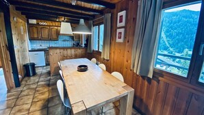 A wooden interior with a dining table, chairs, a kitchen area, and a large window with a view of a forested area. - Chalet Le Jeuna, Morgins (Morgins)