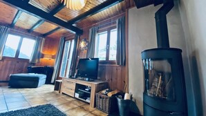 A cozy cabin interior with a wooden ceiling, a television on a wooden stand, a fireplace, and a large window with curtains. - Chalet Le Jeuna, Morgins (Morgins)