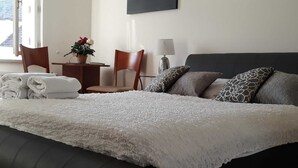 A neatly made bed with a white textured blanket, a dark headboard, and patterned pillows. There is a small table with a plant and a lamp, and two chairs. - Pretty, comfortable apartment (Praga)