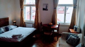 A hotel room with a bed, a table with a bowl of fruit, a chair, a sofa, and two windows with curtains. - Pretty, comfortable apartment (Praga)