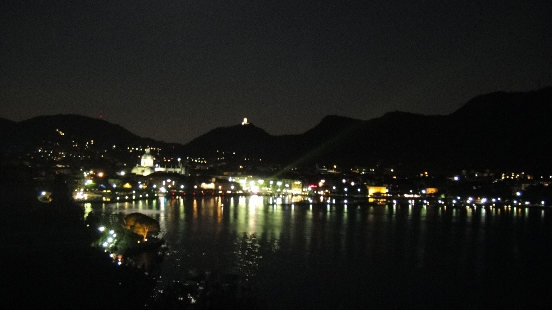 Une vue nocturne d’une ville côtière, avec des bâtiments illuminés dont les reflets se mirent dans l’eau.