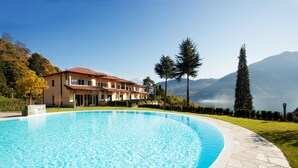 Una piscina con cubierta de piedra, una casa grande de techo rojo y un paisaje montañoso al fondo.