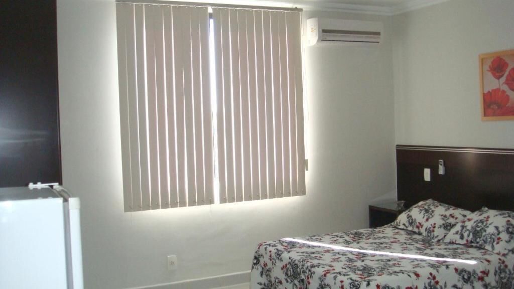 A bedroom with a bed, a nightstand, a window with blinds, and a wall-mounted air conditioner.