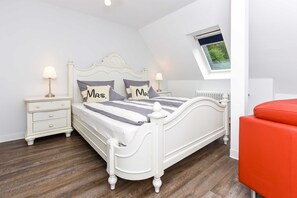 Une chambre à coucher avec un lit blanc, deux tables de chevet avec des lampes, une chaise rouge et une lucarne.