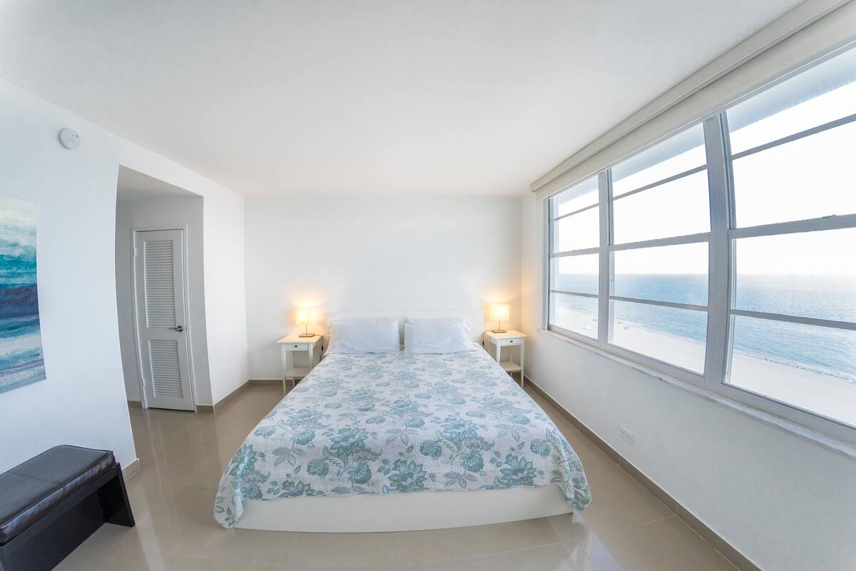 A bedroom with a bed, two bedside tables with lamps, a door, and a large window with a view of the sea.