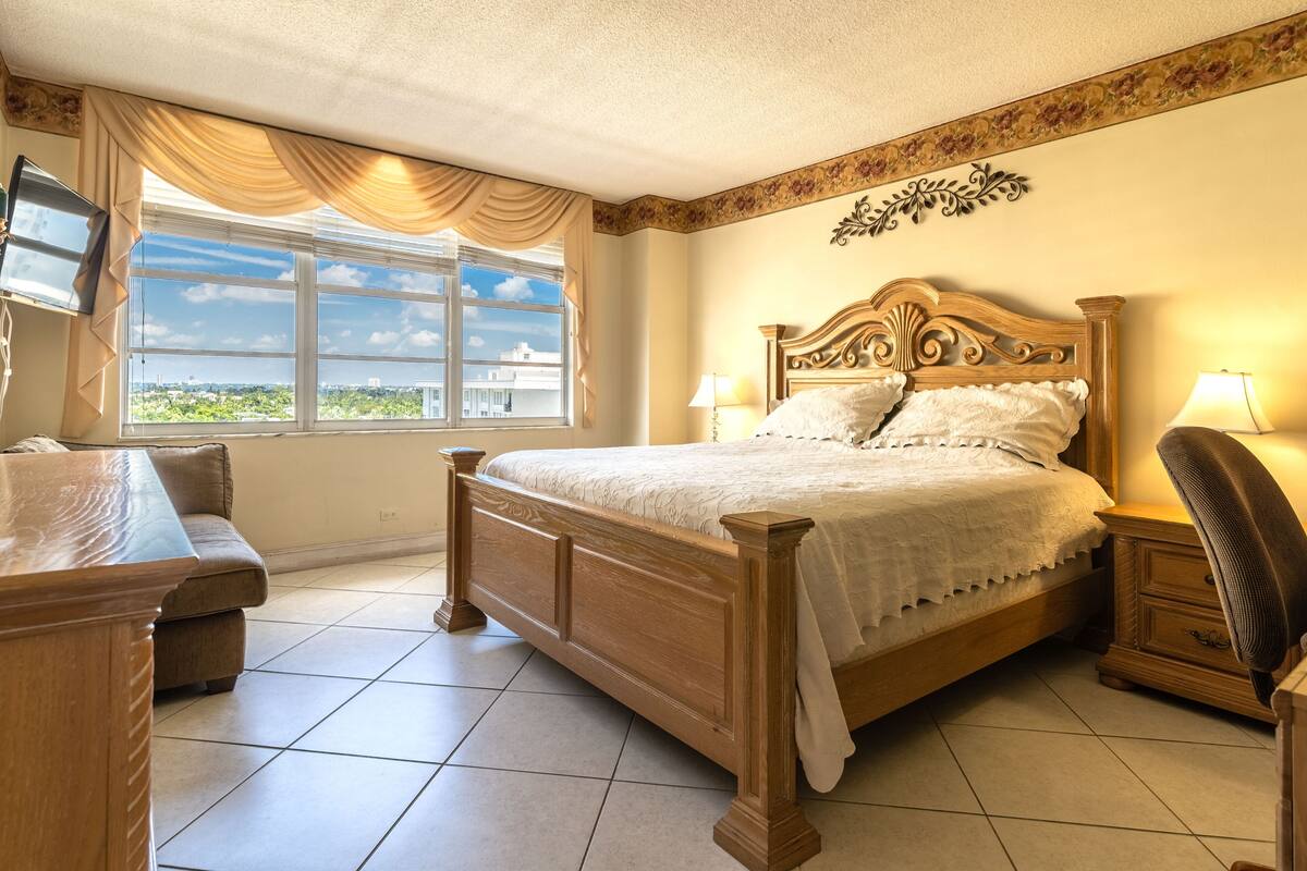 A bedroom with a wooden bed, bedside tables, a nightstand, a chair, and a window with curtains.