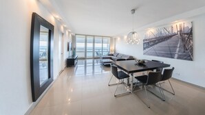 A modern living room with a large window, a dining area with a table and chairs, and a sofa. - Ocean Grand Residences (Miami Beach)