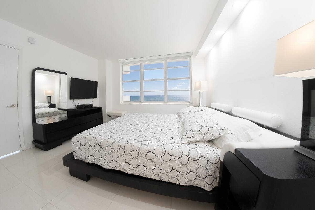 A modern bedroom with a large bed, a TV, a dresser, and a window with a view.