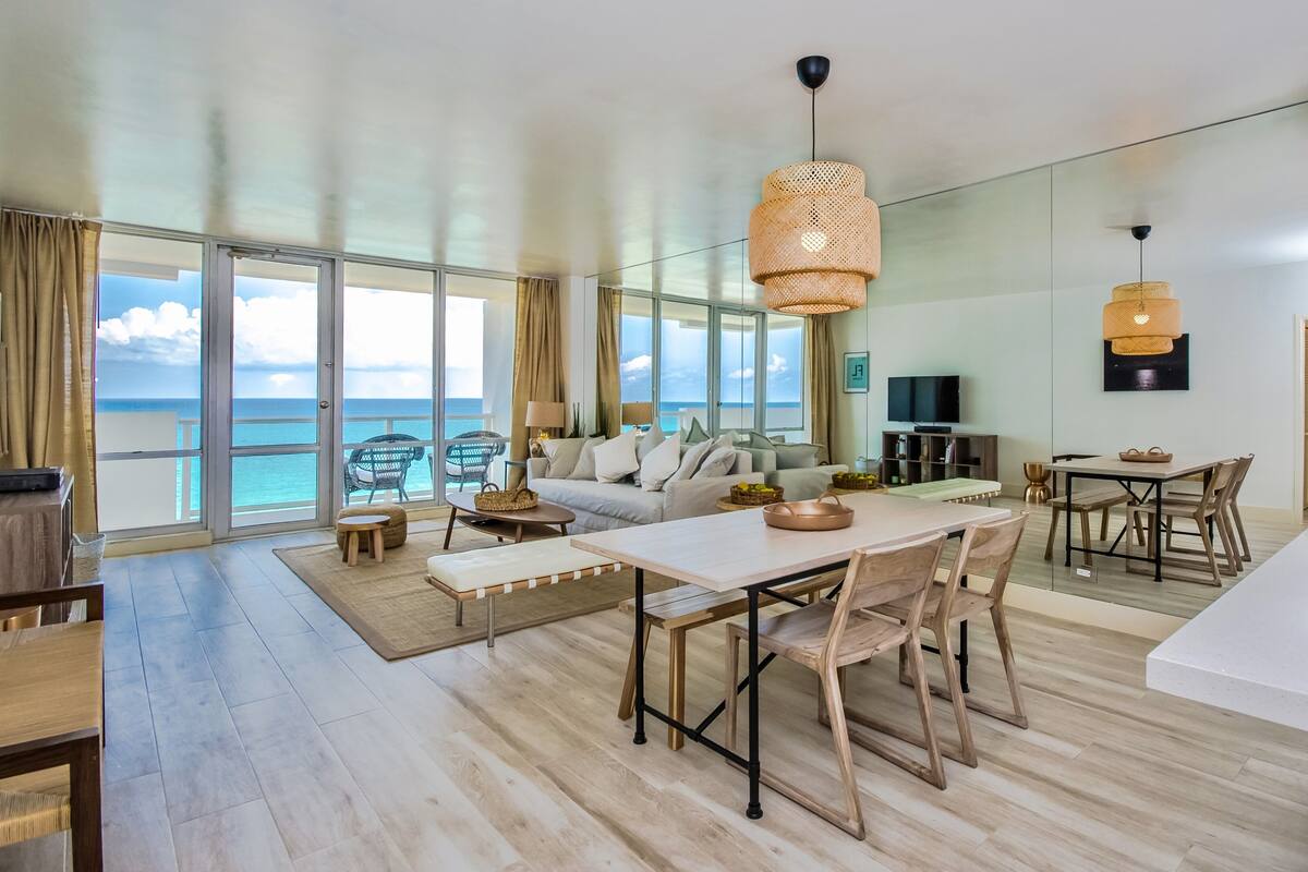 A spacious living area with a large window overlooking the ocean, a dining table with chairs, a sofa, and a television.