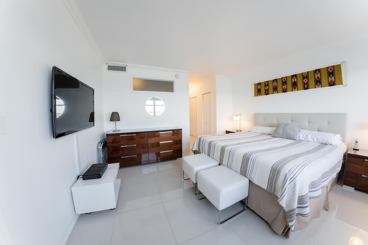 A modern bedroom with a large bed, bedside tables, a TV mounted on the wall, a dresser, and a small desk with a lamp.