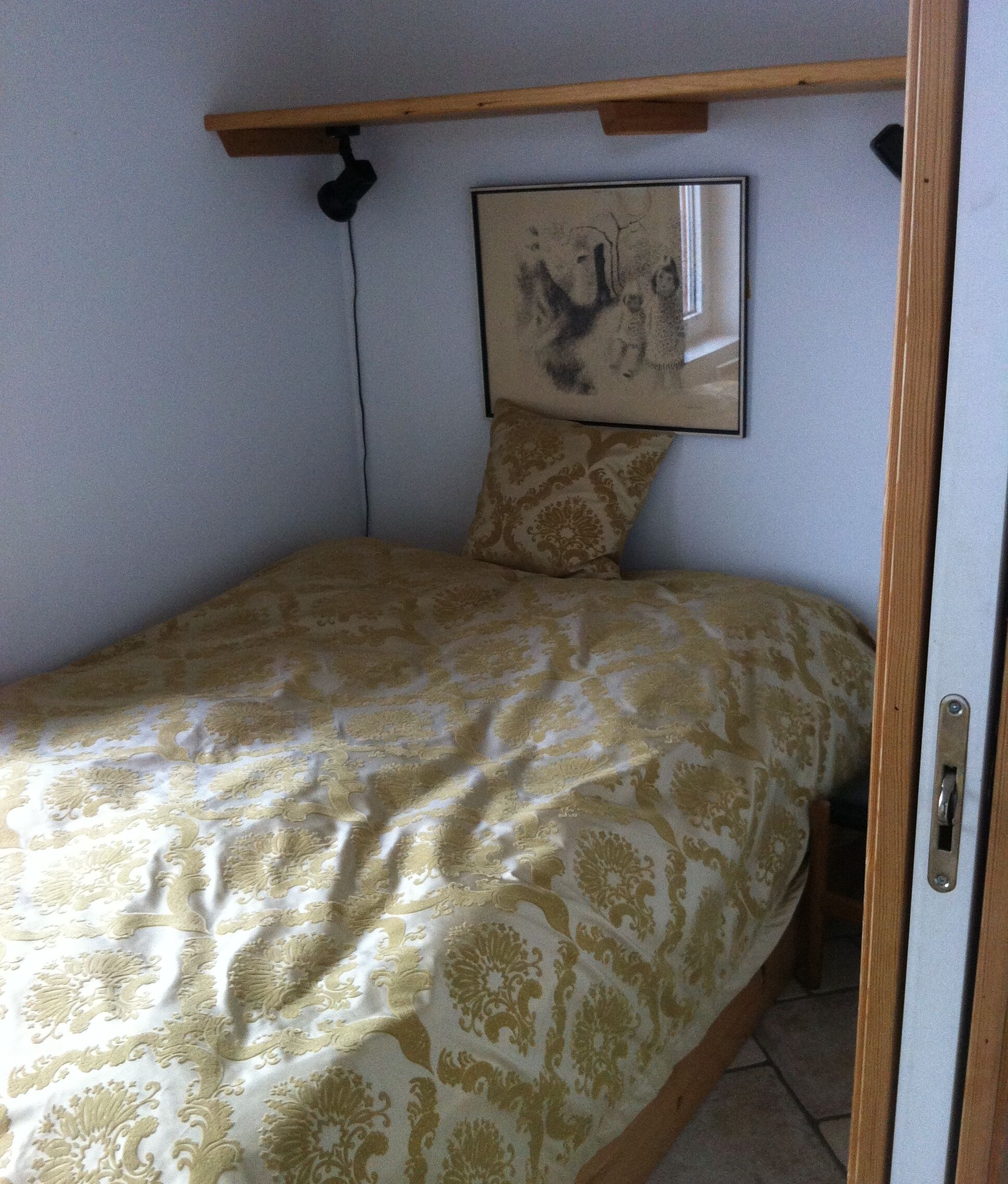 Une petite chambre avec un lit simple, une couvre-lit à motifs, une tête de lit en bois et un tableau encadré au mur.