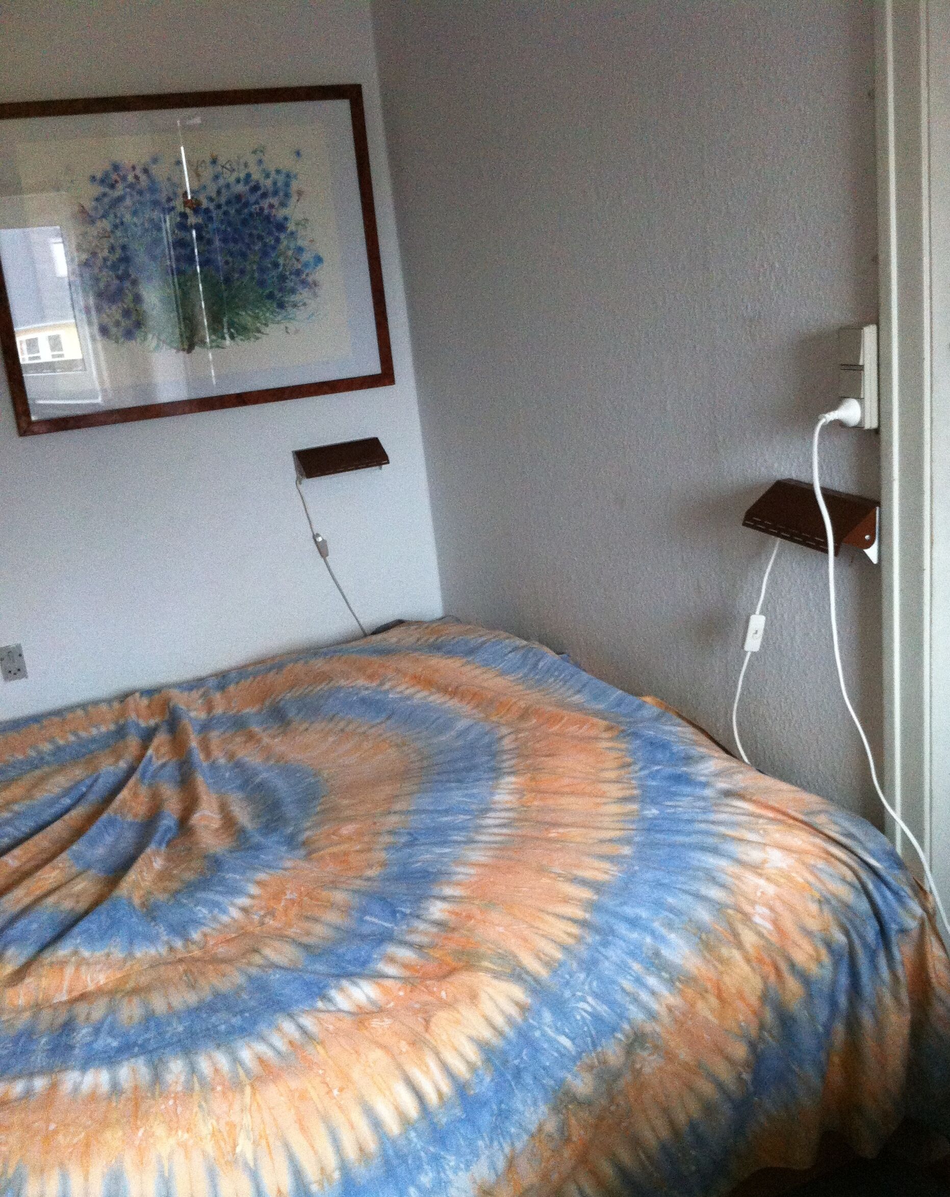 A bedroom with a bed covered in a tie-dye blanket, a framed picture on the wall, and a power outlet with a charging cable.