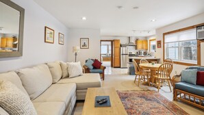A spacious living area with a sofa, a coffee table, and a dining table with chairs. There is a kitchen in the background with wooden cabinets and a refrigerator.