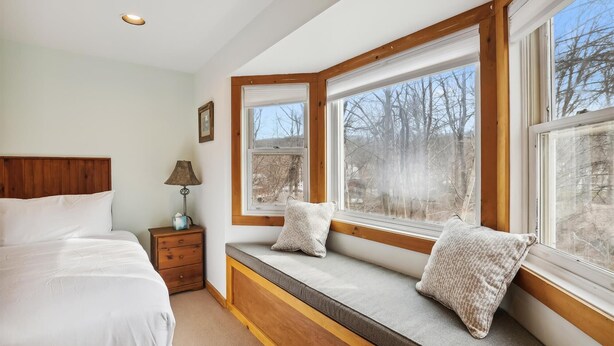 A bedroom with a large window, a bedside table with a lamp, and a bed with pillows.