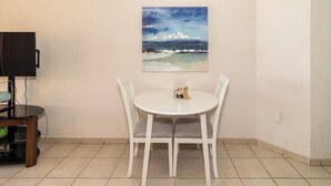 Un coin repas avec une table et des chaises blanches, un tableau représentant une scène de plage et un téléviseur posé sur un meuble.