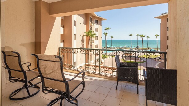Un balcon avec deux fauteuils à bascule et une table, donnant sur une plage bordée de palmiers et offrant une vue dégagée sur l’océan.