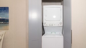Une petite buanderie fermée, comprenant un lave-linge et un sèche-linge, une petite chaise et un tableau accroché au mur.