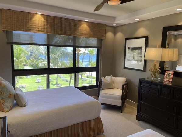 Un dormitorio con cama, una silla, cómoda, ventana con vista a un jardín y palmeras, y ventilador de techo.