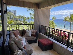 Un balcon avec un canapé et un pouf, donnant sur une plage bordée de palmiers et offrant une vue sur un ciel dégagé.