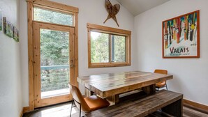 Un comedor con mesa de madera y bancos, ventanas amplias y una obra de arte colgada en la pared.