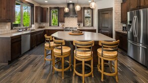A modern kitchen with a central island, dark wooden cabinets, stainless steel appliances, and a large window offering a view of greenery.
