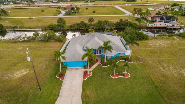 An aerial view of a house with a blue garage, surrounded by a well-maintained lawn, a small pool, and a dock extending into a body of water.
