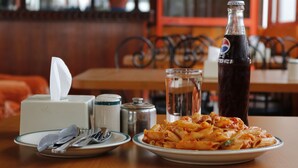 Une table à manger avec une assiette de pâtes, une bouteille de soda, un verre d’eau, un salier et un porte-serviettes.
