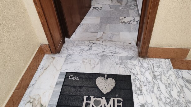 Un pasillo con suelo de mármol que conduce a una puerta. Un felpudo con la inscripción 'Geko HOME' se encuentra en la entrada.