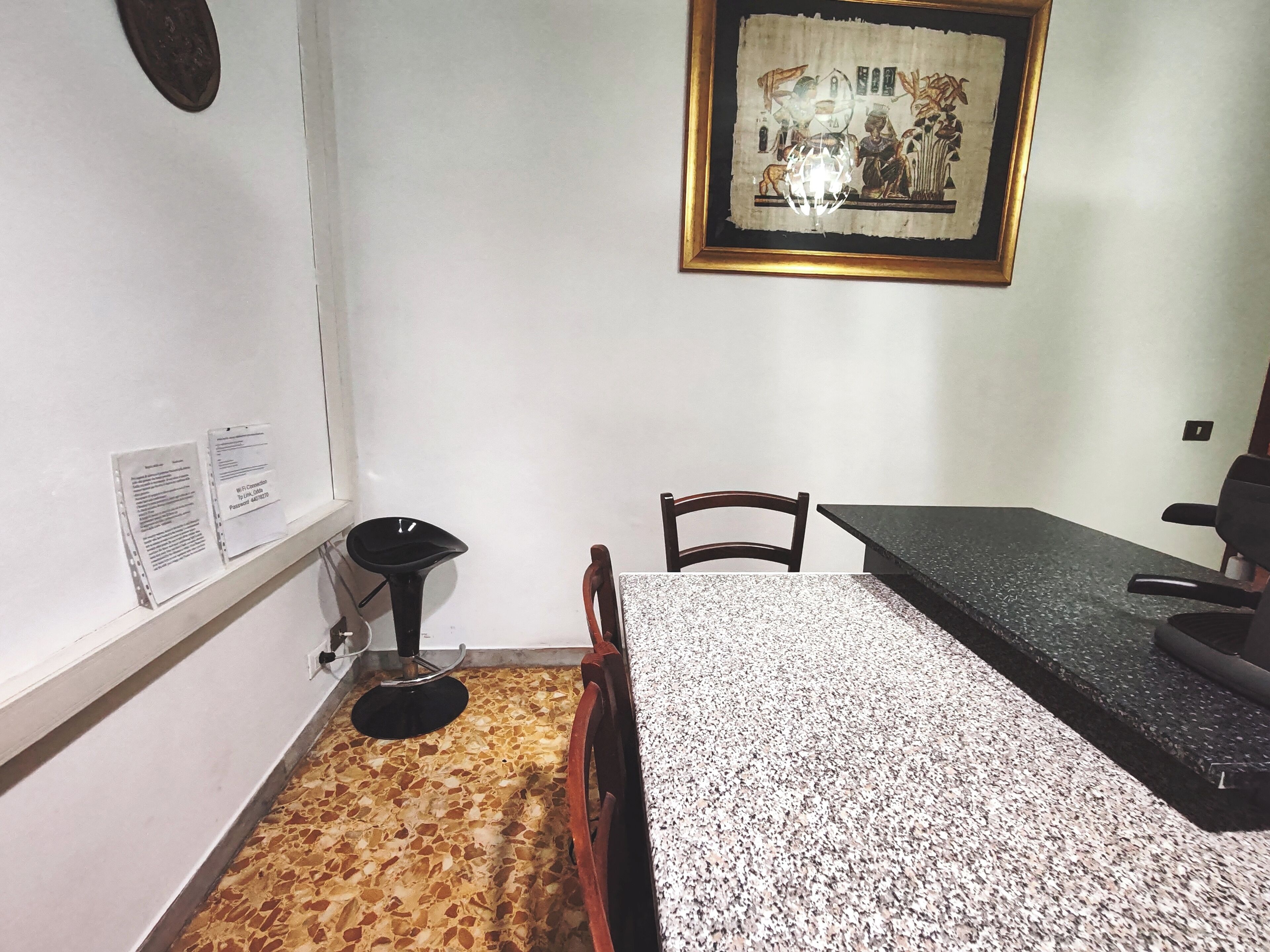 Un petit coin repas avec une table, des chaises, une chaise noire, un tableau encadré au mur et un sol carrelé.