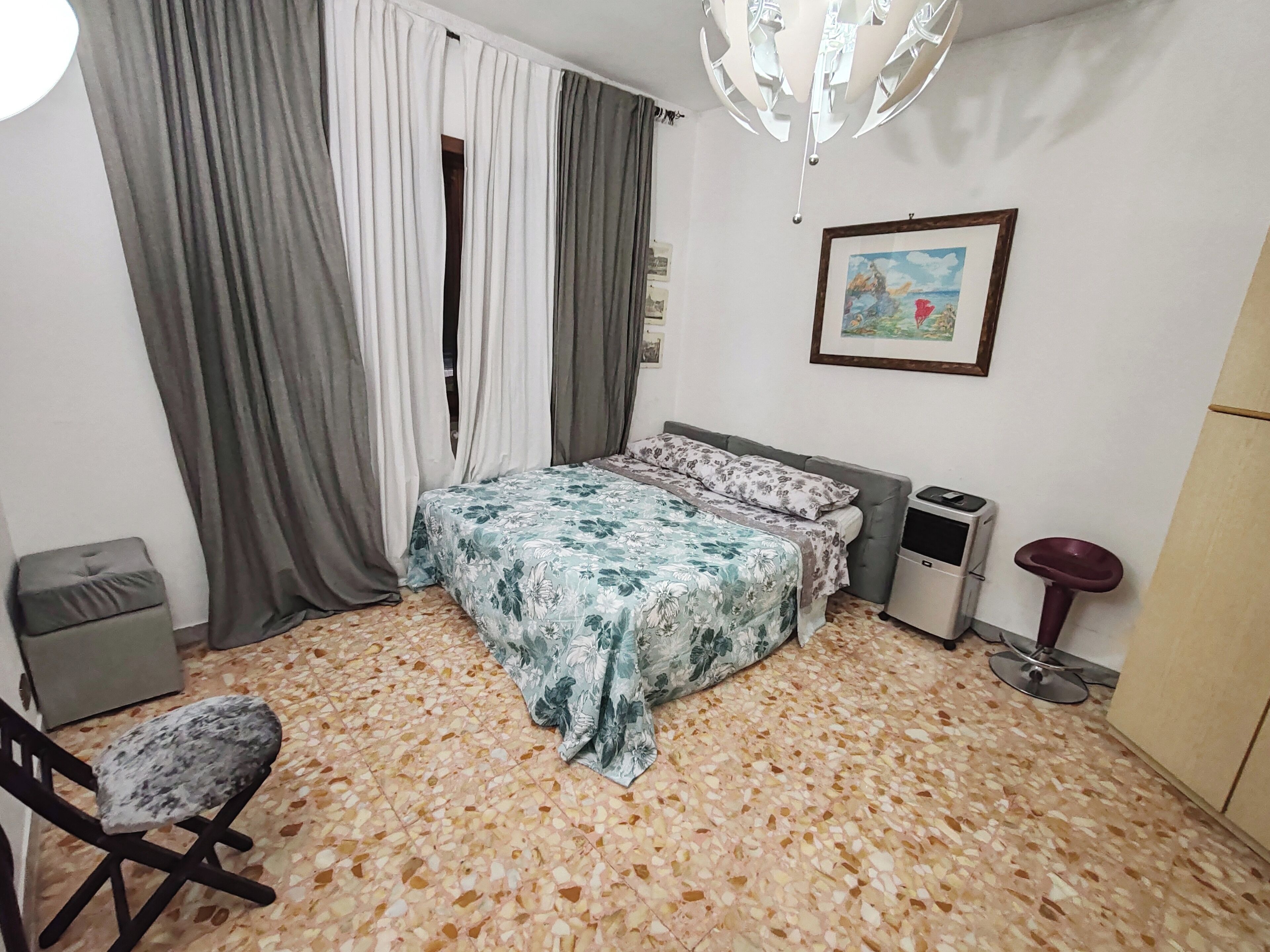 A bedroom with a bed, a chair, a small table, a waste bin, and a picture on the wall.