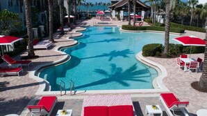A swimming pool with red and white lounge chairs and umbrellas, surrounded by palm trees and a building with a tiled roof.