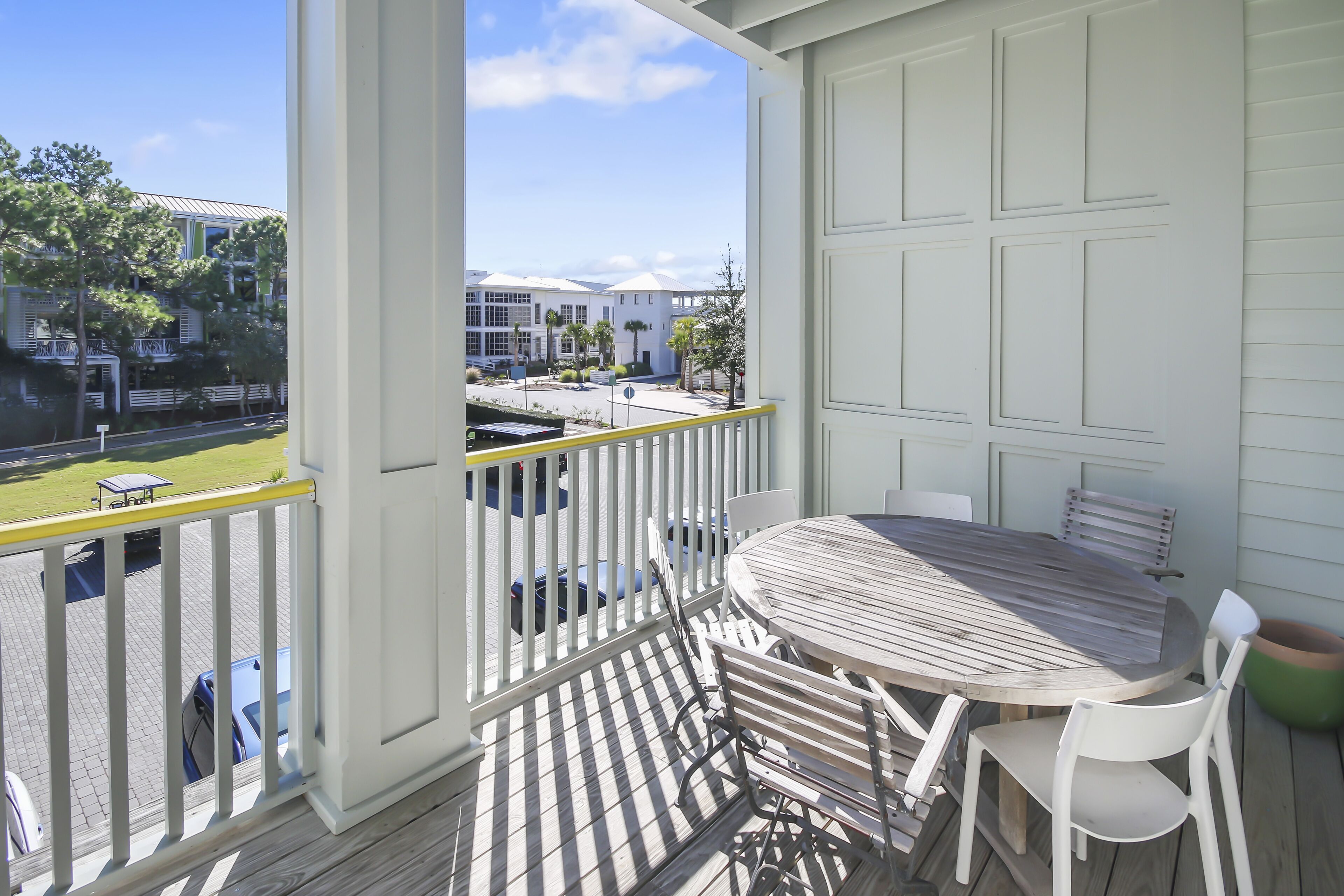 A balcony with a wooden table and chairs, a white railing, and a view of a parking area and buildings in the background.