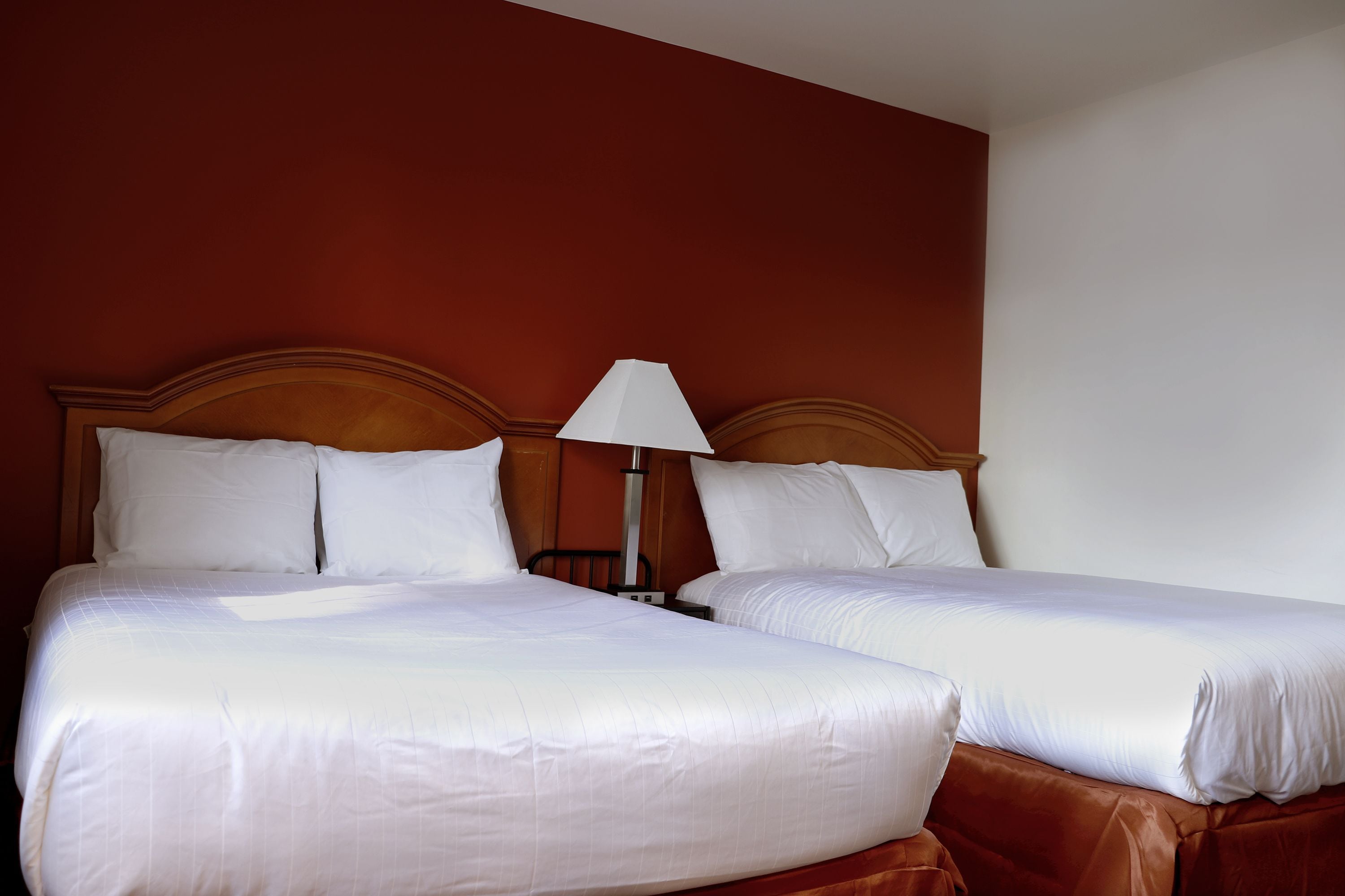 Una cama doble con ropa blanca y un cabecero de madera, una mesita de noche con lámpara, y una pared de acento roja.