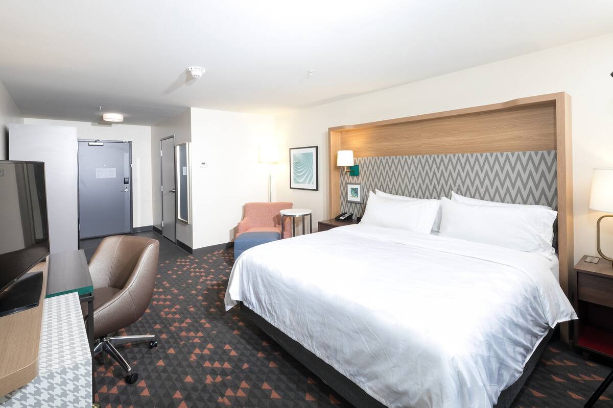 A hotel room with a large bed, a desk with a computer, a chair, a small table, and a nightstand with a lamp.