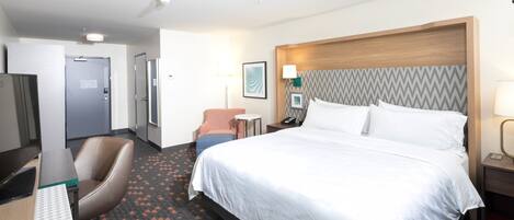 A hotel room with a large bed, a desk with a computer, a chair, a small table, and a nightstand with a lamp.