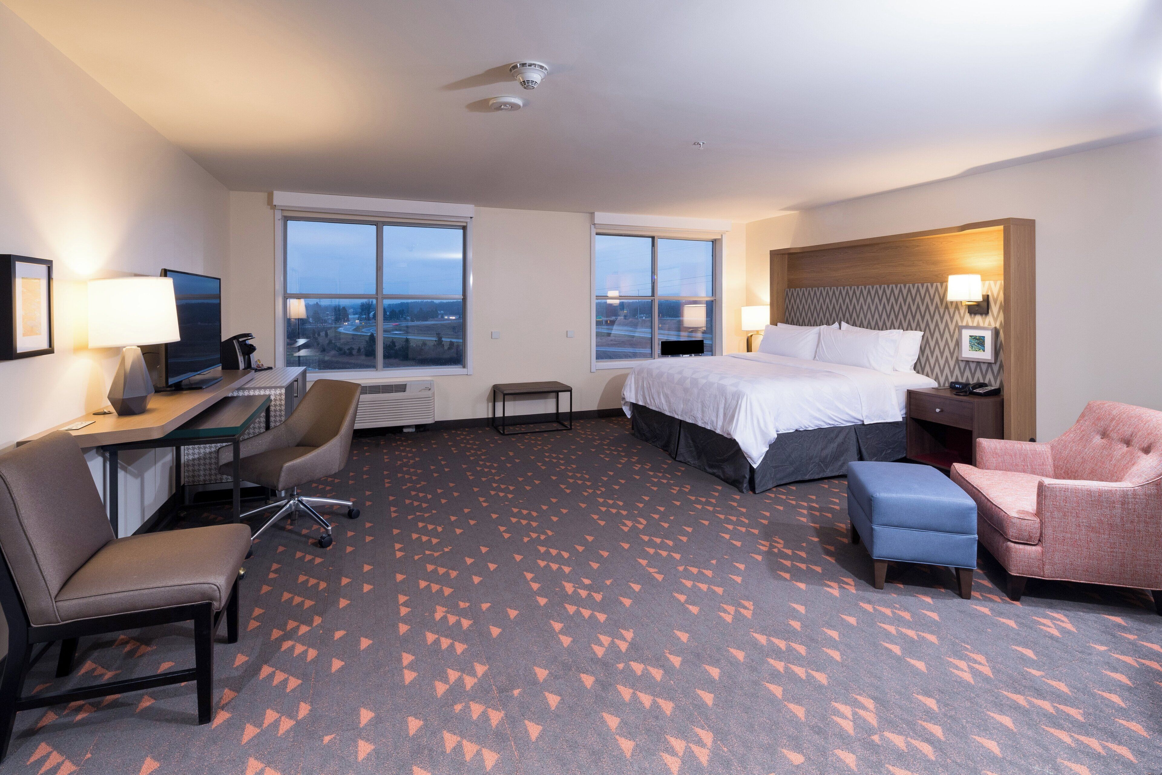 A hotel room with a bed, desk, chair, and a view of the city through large windows.